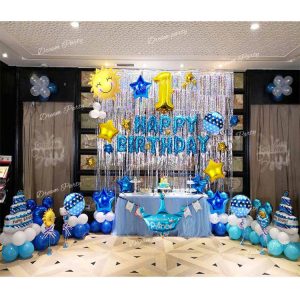 A colorful birthday setup with a blue "Happy Birthday" foil balloon, silver curtains, a gold digit balloon, sun and cake balloons, a crown balloon, star balloons, and blue, white, and light blue latex balloons.
